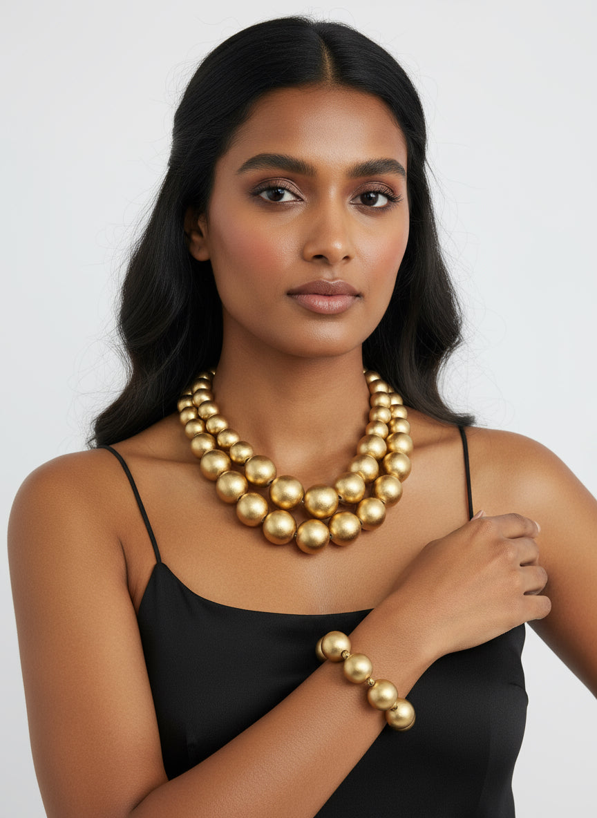 Gold necklace on a black stand with a white box and green flowers against a brick wall.