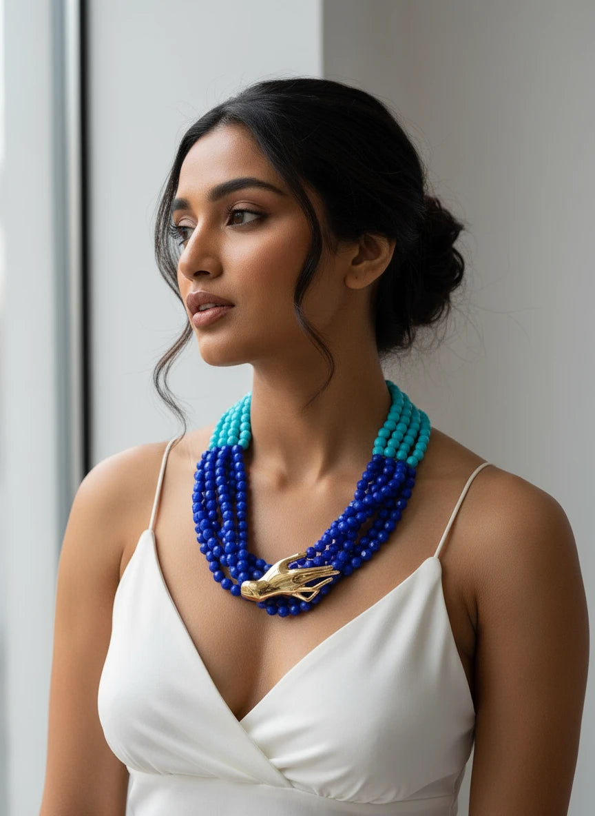 Blue beaded necklace with a gold hand pendant on a black stand.