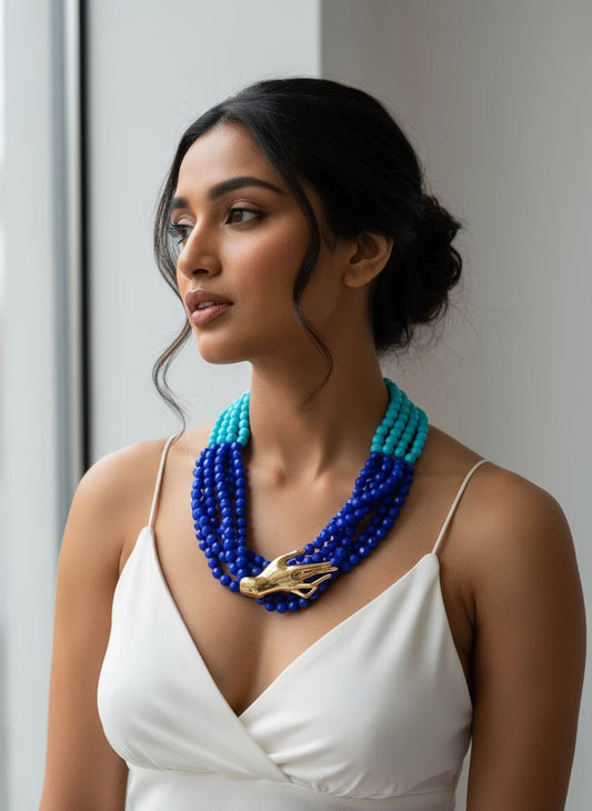 Blue beaded necklace with a gold hand pendant on a black stand.